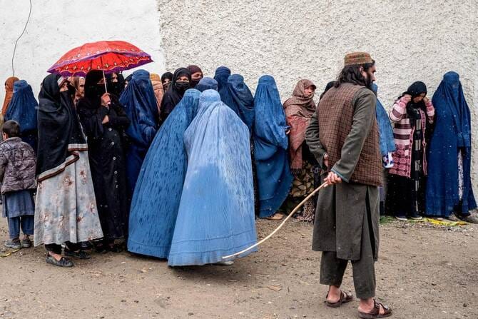 TOPSHOT - A Taliban security personnel stands guard as Afghan burqa-clad women wait in queue in the midst of a downpour to receive food supplies donated during the Islamic holy fasting month of Ramadan, in Kabul on March 25, 2025 (AFP)