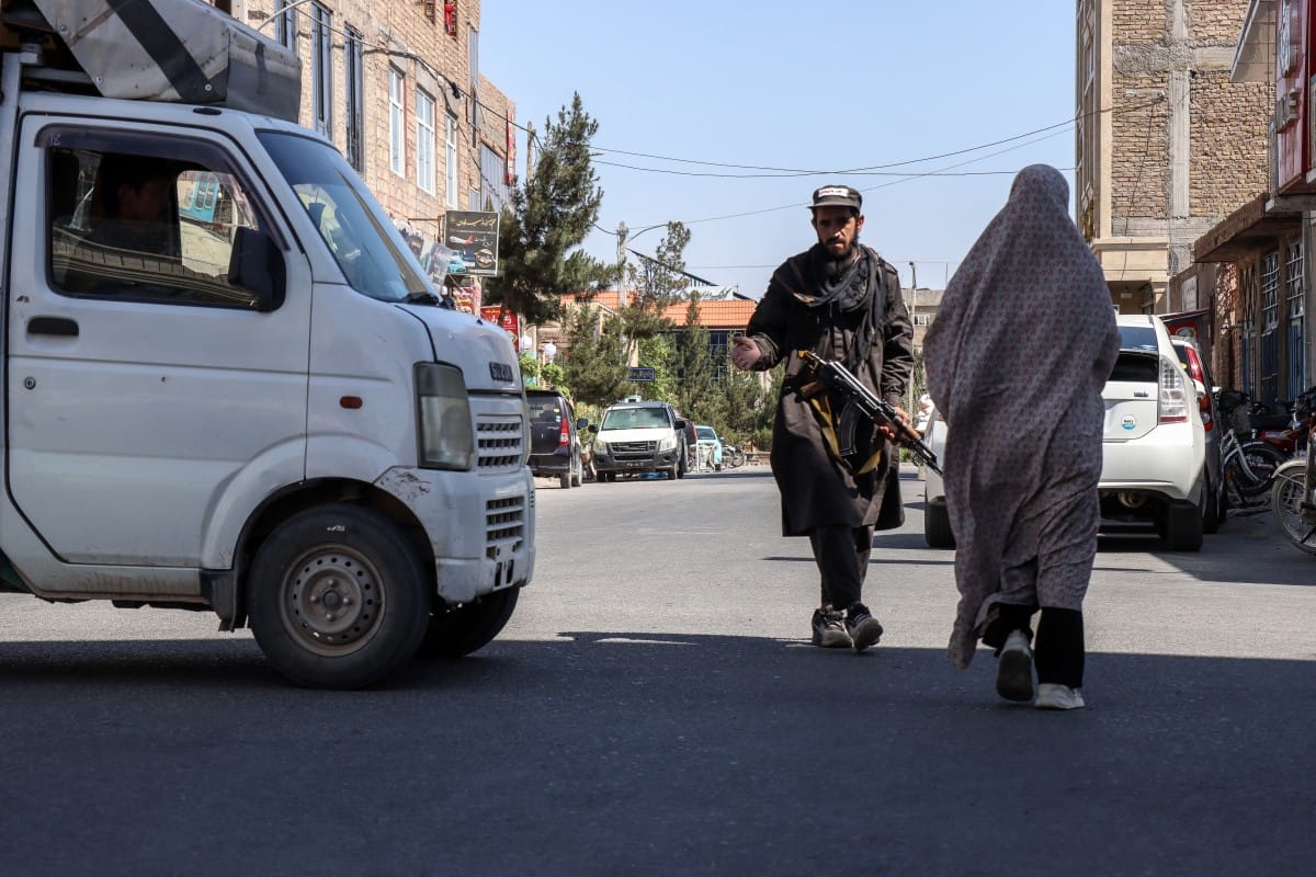 An Afghan burqa-clad woman walks past a Taliban security personnel standing guard outside a mosque in Shahrak-e-Almahdi, Jebrail district of Herat Province on June 11, 2026 (AFP)