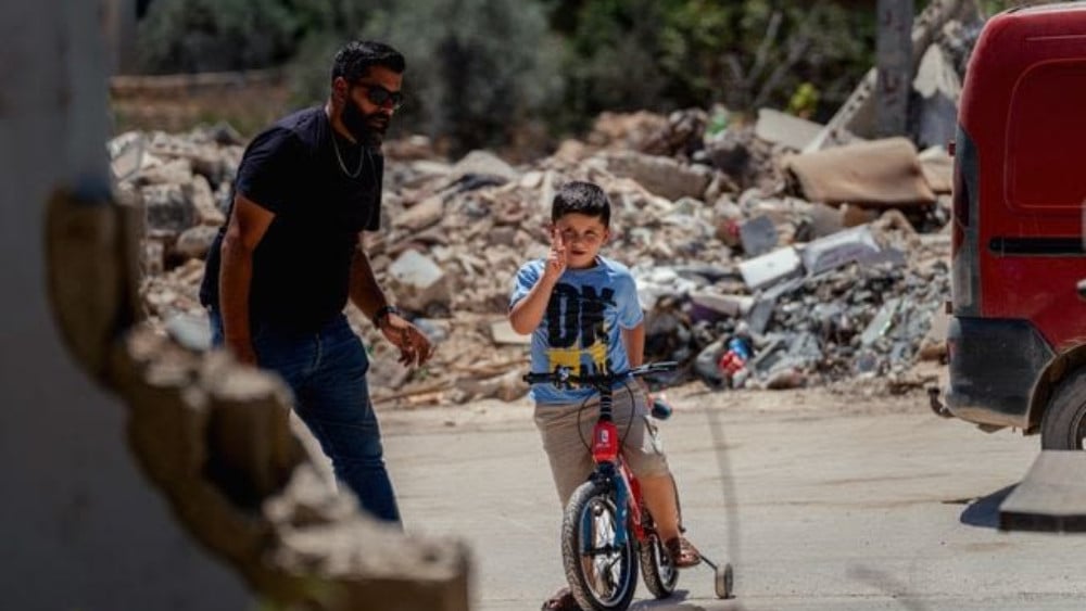 A child rides a bicycle as a man walks beside him through a neighborhood covered in piles of rubble and debris in Maifadoun, southern Lebanon, on July 31, 2026. (AFP)