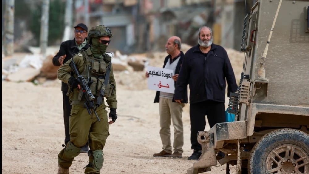 An Israeli soldier walks near Palestinians protesting after Israeli security forces blocked the entrance of the Nur Shams Palestinian refugee camp in the occupied West Bank on February 9, 2026, demanding to be allowed to return to their homes from where t