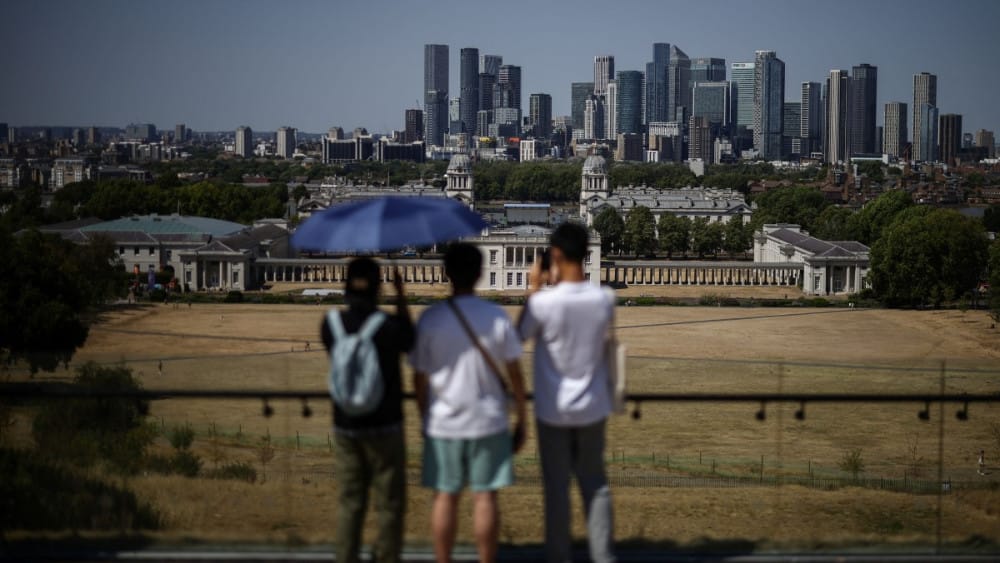 People shelter from the sun as they look out at the Old Royal Naval College (behind C) and the skyline of Canary Wharf, across the dried out and yellow grass in Greenwich Park, south-east London on July 29, 2026. (AFP)