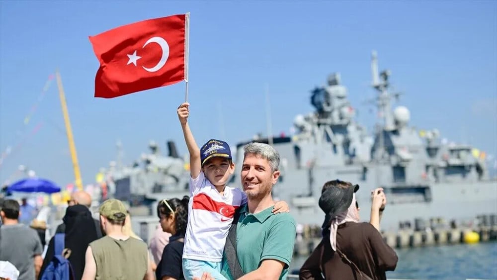 A child holds a Turkish flag as he poses with a man in front of a Turkish Navy vessel during TEKNOFEST Blue Homeland at the Golcuk Naval Shipyard Command in Kocaeli, Turkiye, on August 23, 2026. (AFP)
